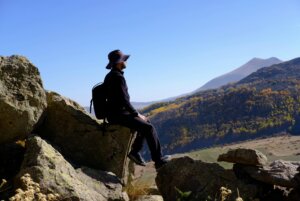 Man in black hiking outfit and hat sitting on rocks overlooking a mountainous landscape with autumn foliage.