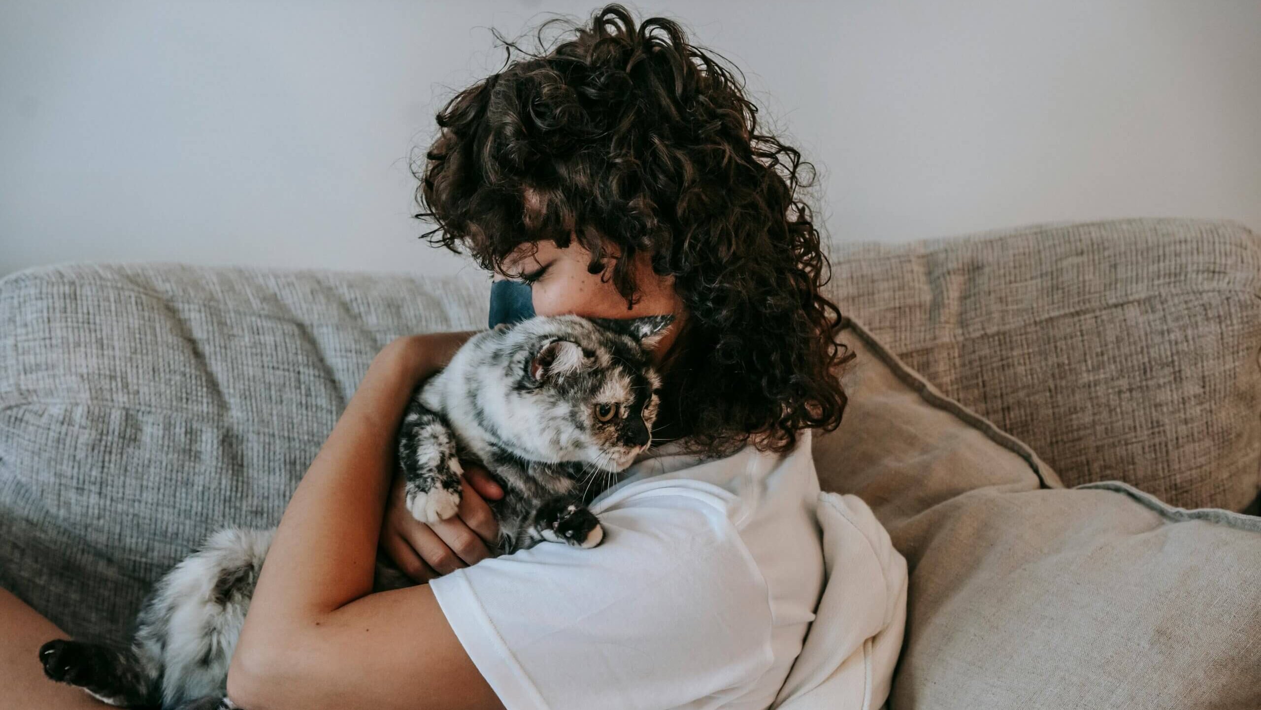 Person with curly hair hugging a gray and black cat on a beige couch with cushions