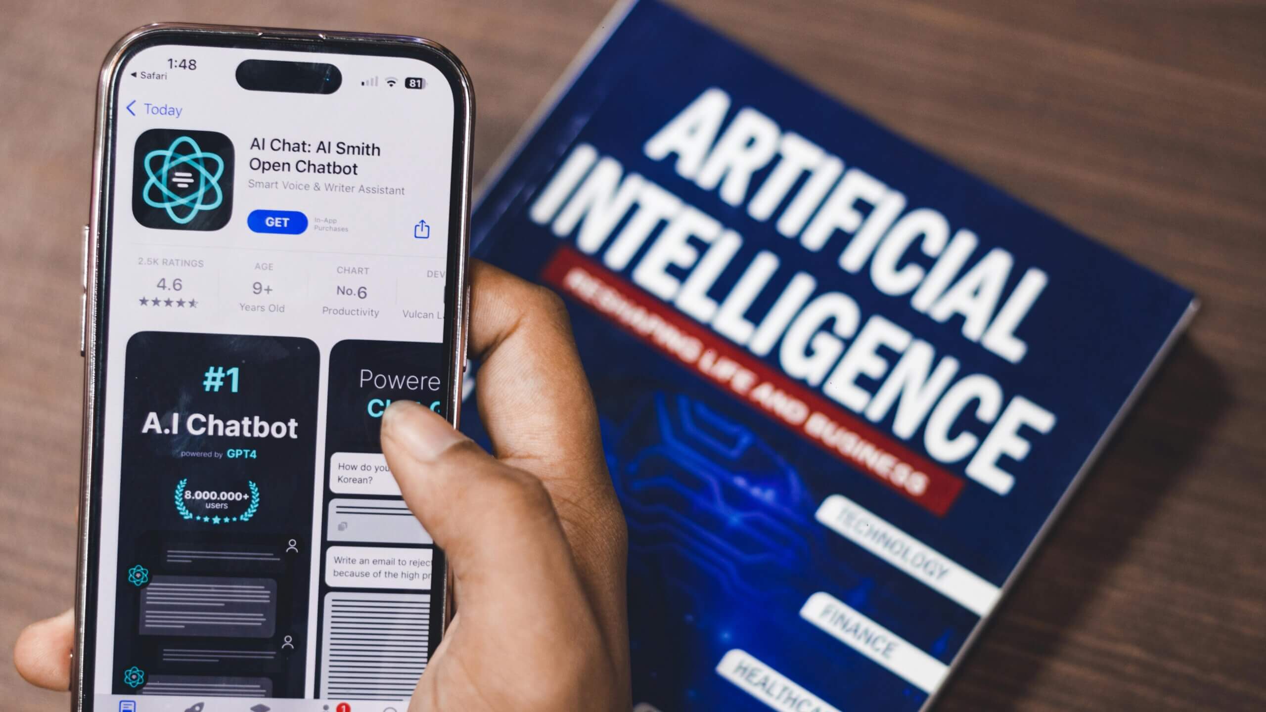 Hand holding iPhone displaying AI Chat: AI Smith Open Chatbot app next to a book titled "Artificial Intelligence"