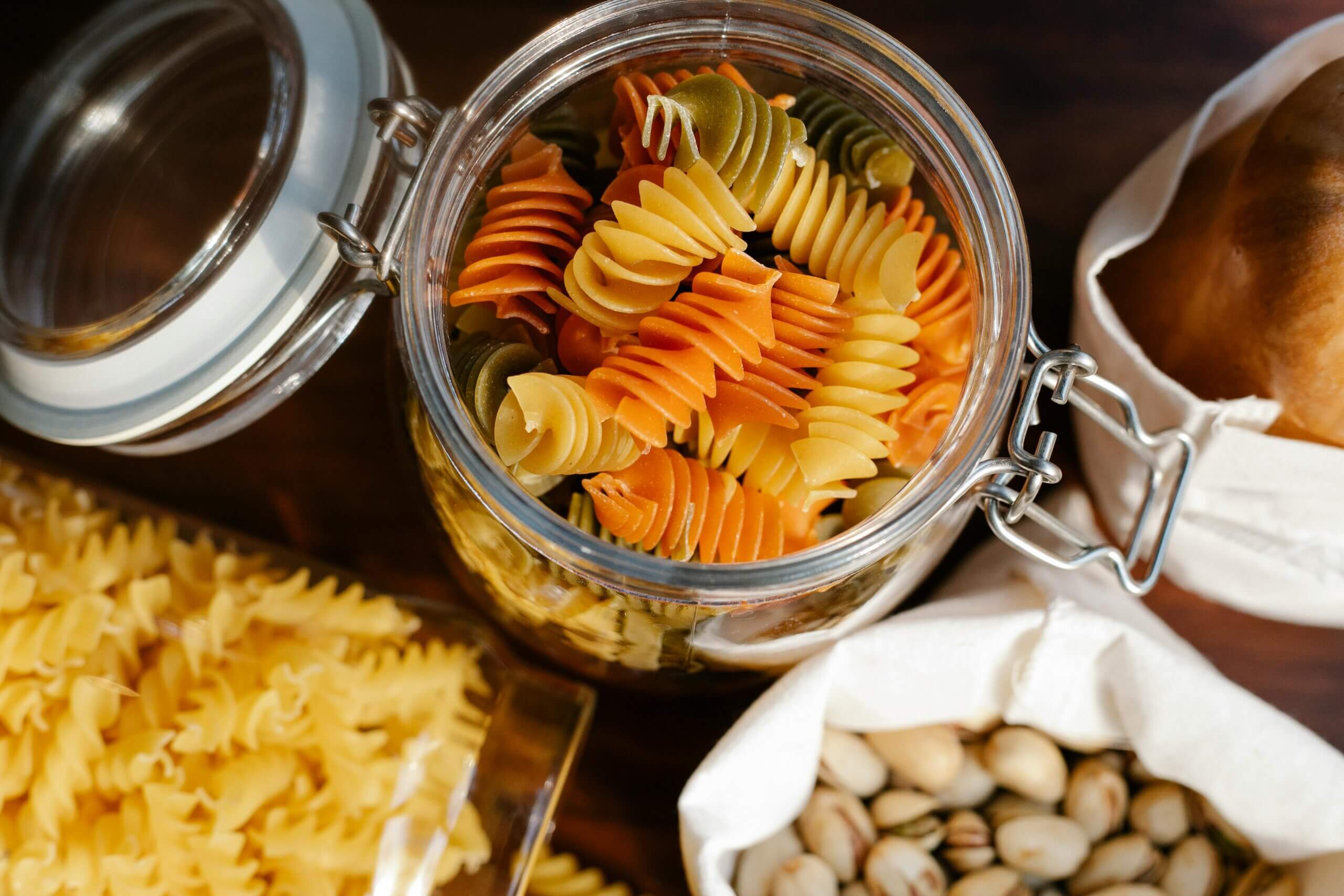 Glass jar filled with tri-color rotini pasta surrounded by yellow pasta, pistachios, and bread in cloth bags