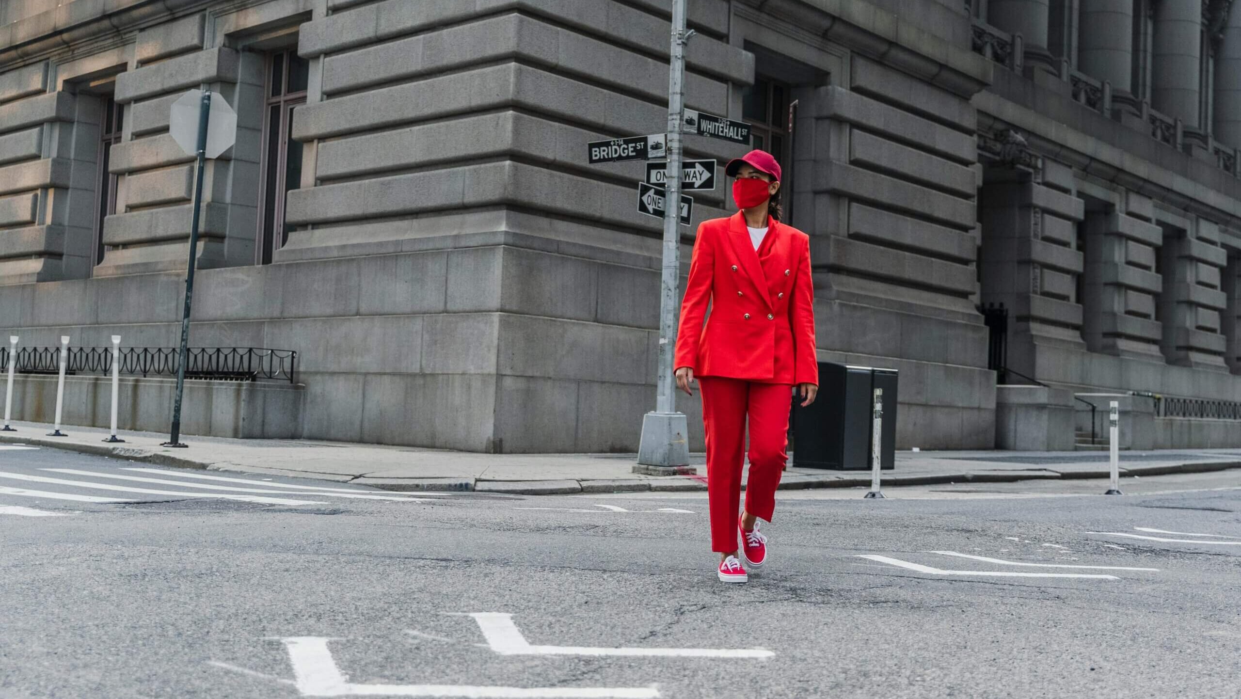 Person in red suit, red cap, and red mask walking on a city street corner near Bridge St and Whitehall St signs