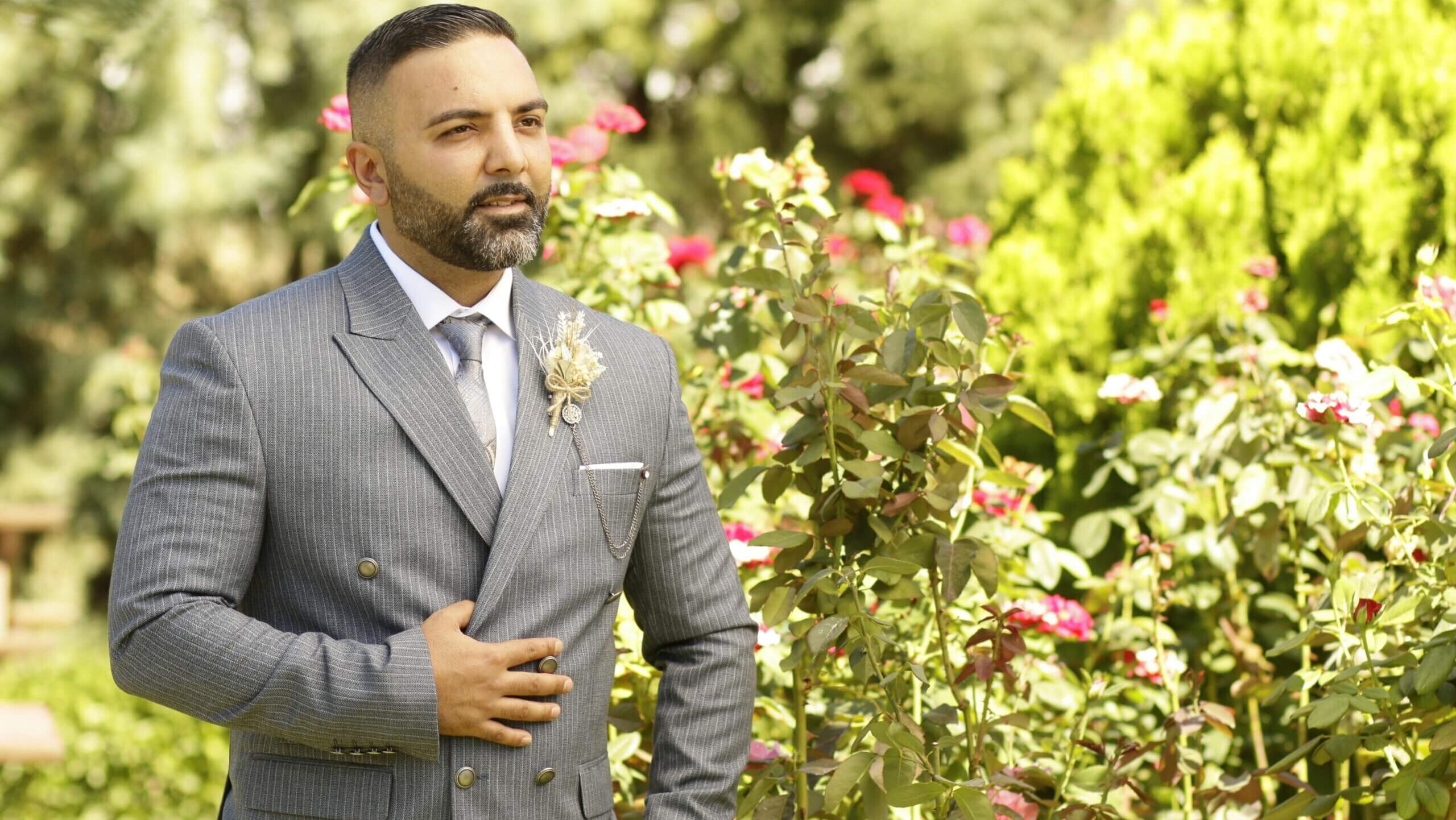 Man in a gray pinstripe suit with boutonniere standing beside blooming rose bushes in a garden.
