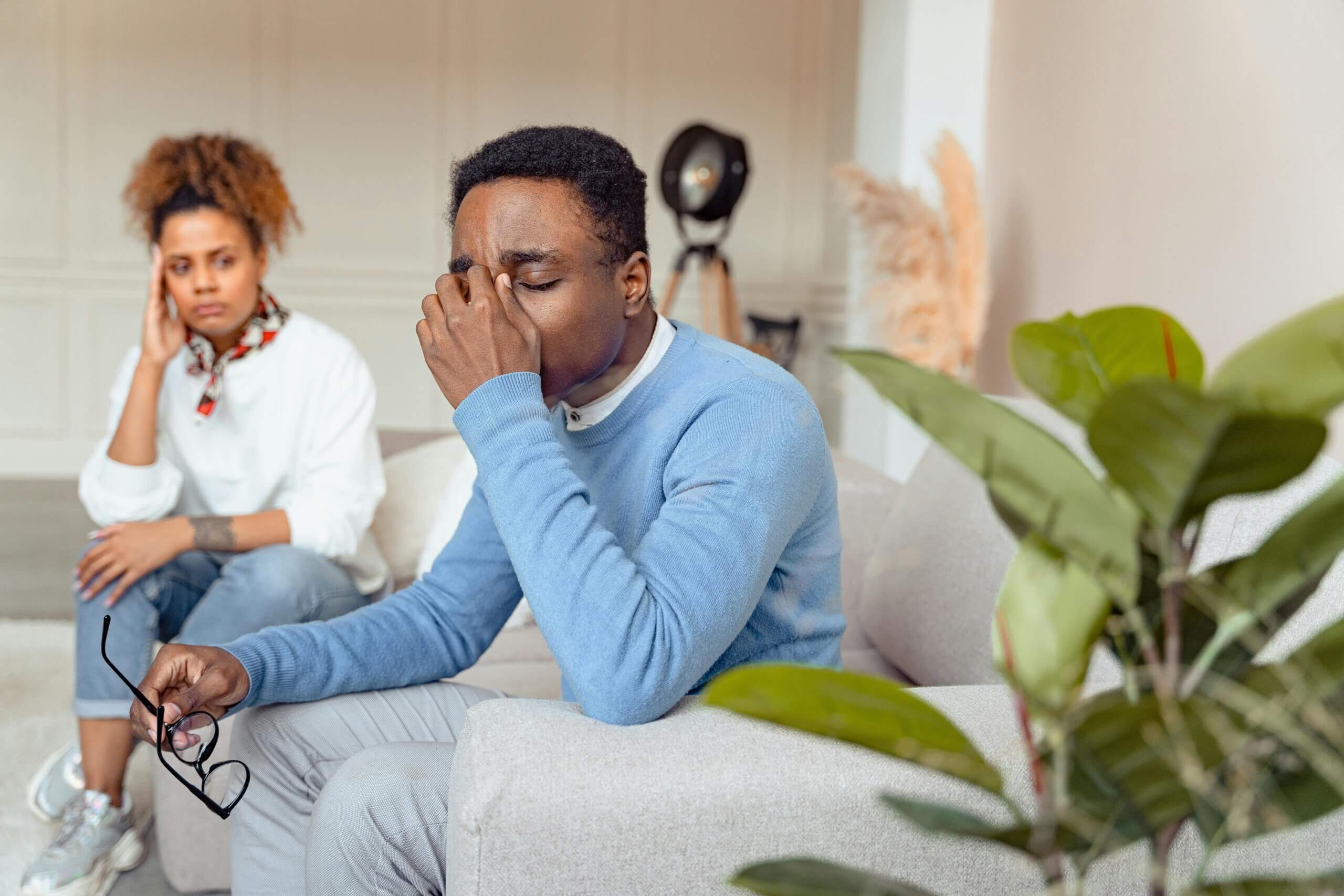 Man in blue sweater holding glasses and rubbing eyes while woman in white shirt watches concerned on couch