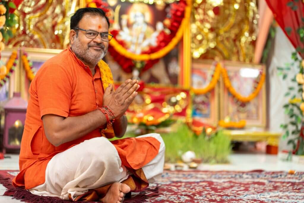 Man in orange kurta and white dhoti sitting cross-legged with hands in namaste gesture in decorated prayer room