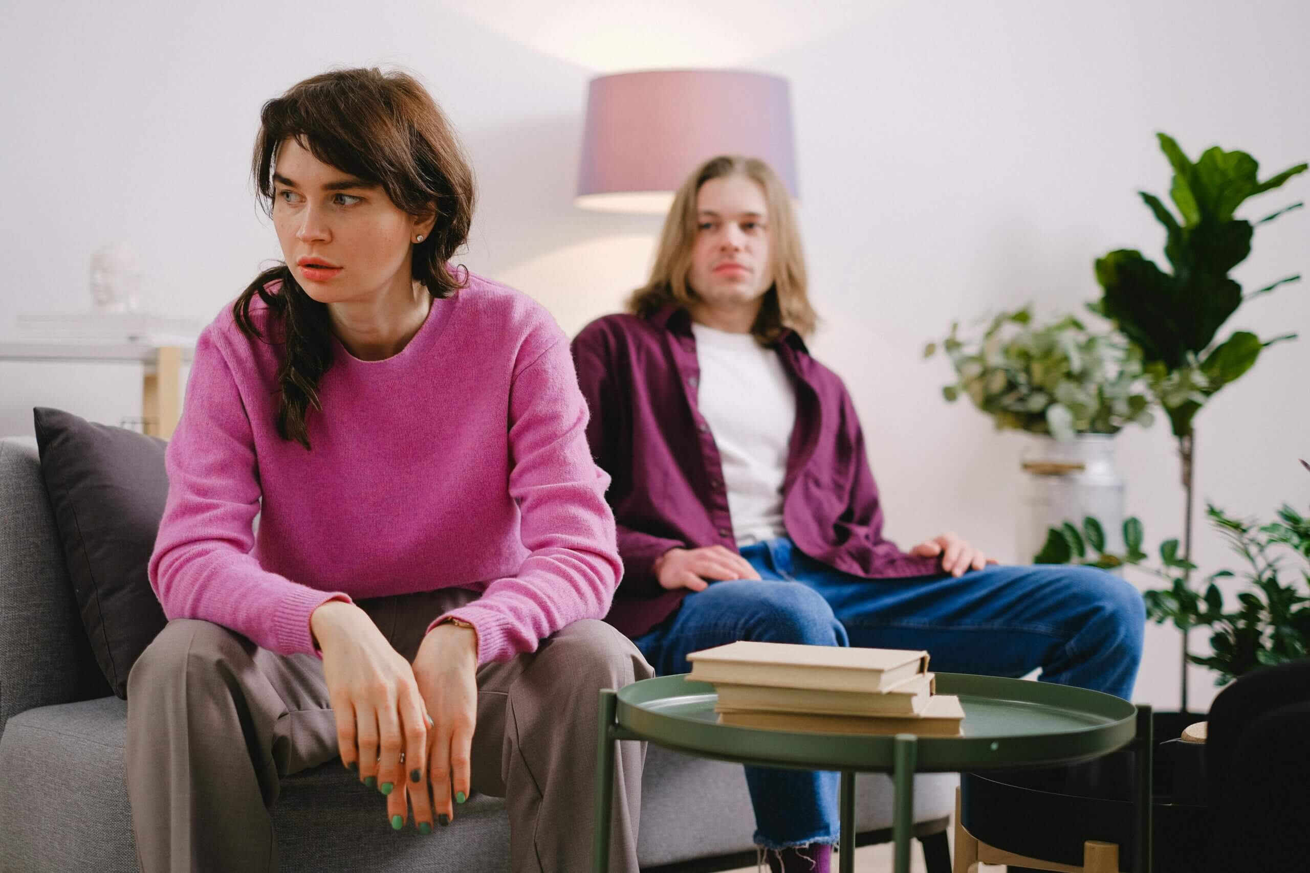 Two young adults sitting on a couch looking upset, with a green table and books in front of them.