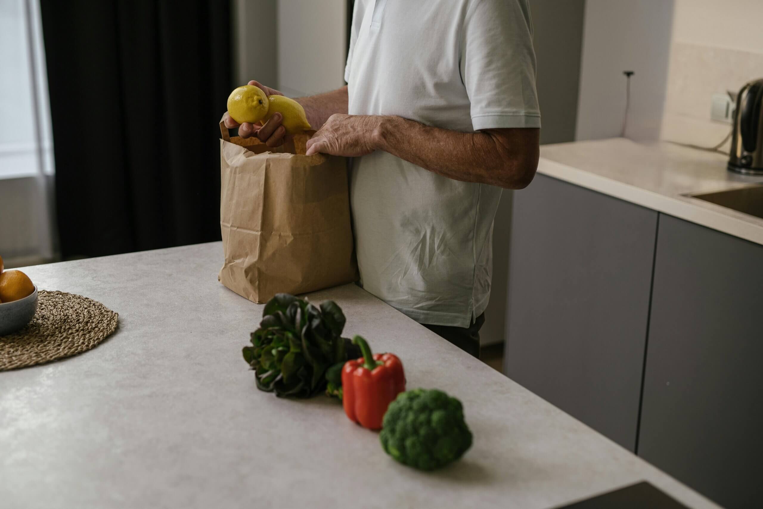 Person unpacking lemons from a paper grocery bag in a modern kitchen with vegetables on the counter