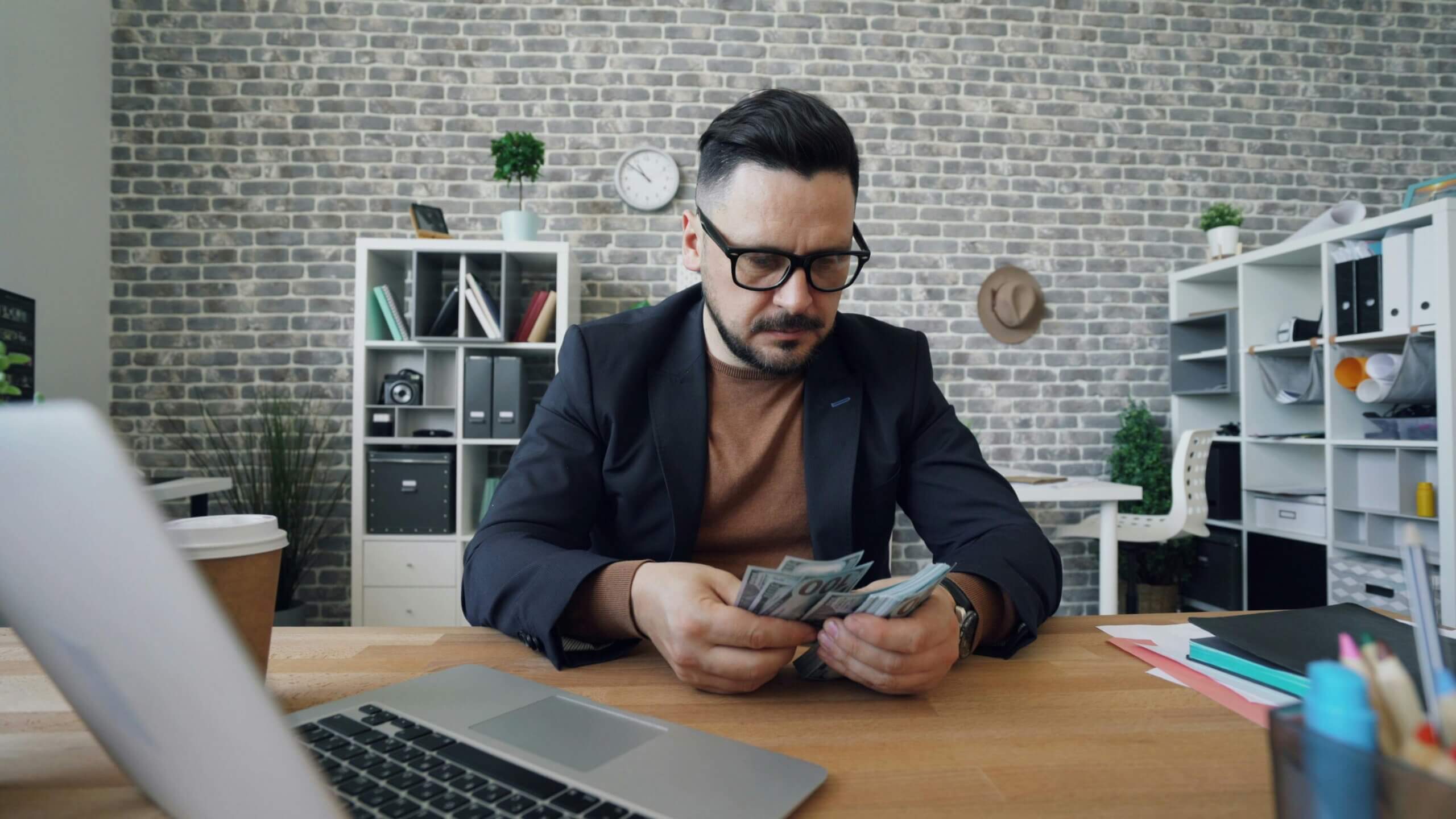 Man in glasses and blazer counting US dollar bills at office desk with laptop and coffee cup