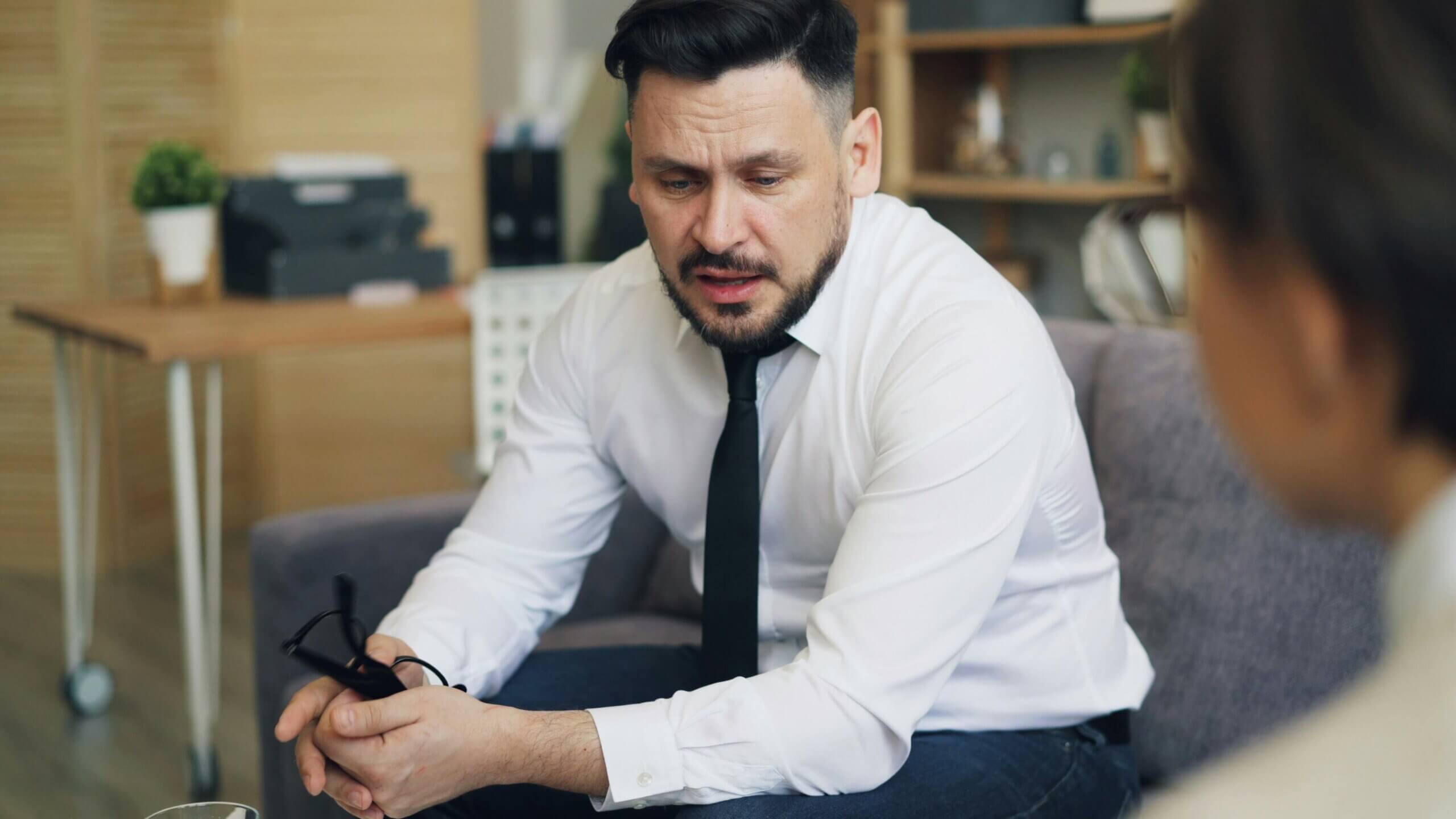 Man in white shirt and black tie holding glasses, appearing distressed during a conversation in an office setting