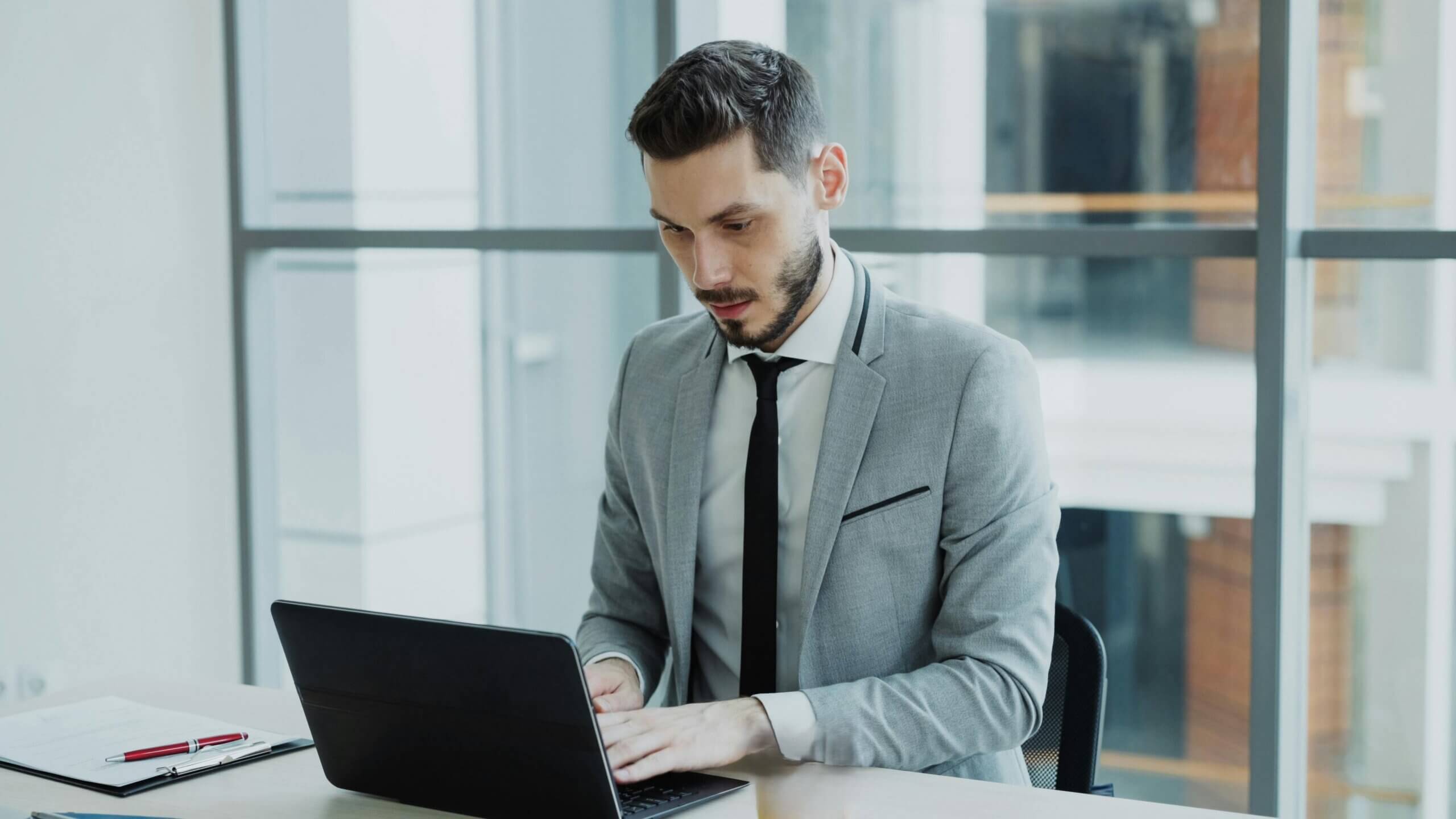 Man in gray suit typing on laptop at office desk with clipboard and pen nearby