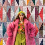 Person with rainbow hair and makeup wearing a pink fur coat, green beret, and colorful blouse against a geometric mosaic wall