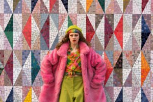 Person with rainbow hair and makeup wearing a pink fur coat, green beret, and colorful blouse against a geometric mosaic wall