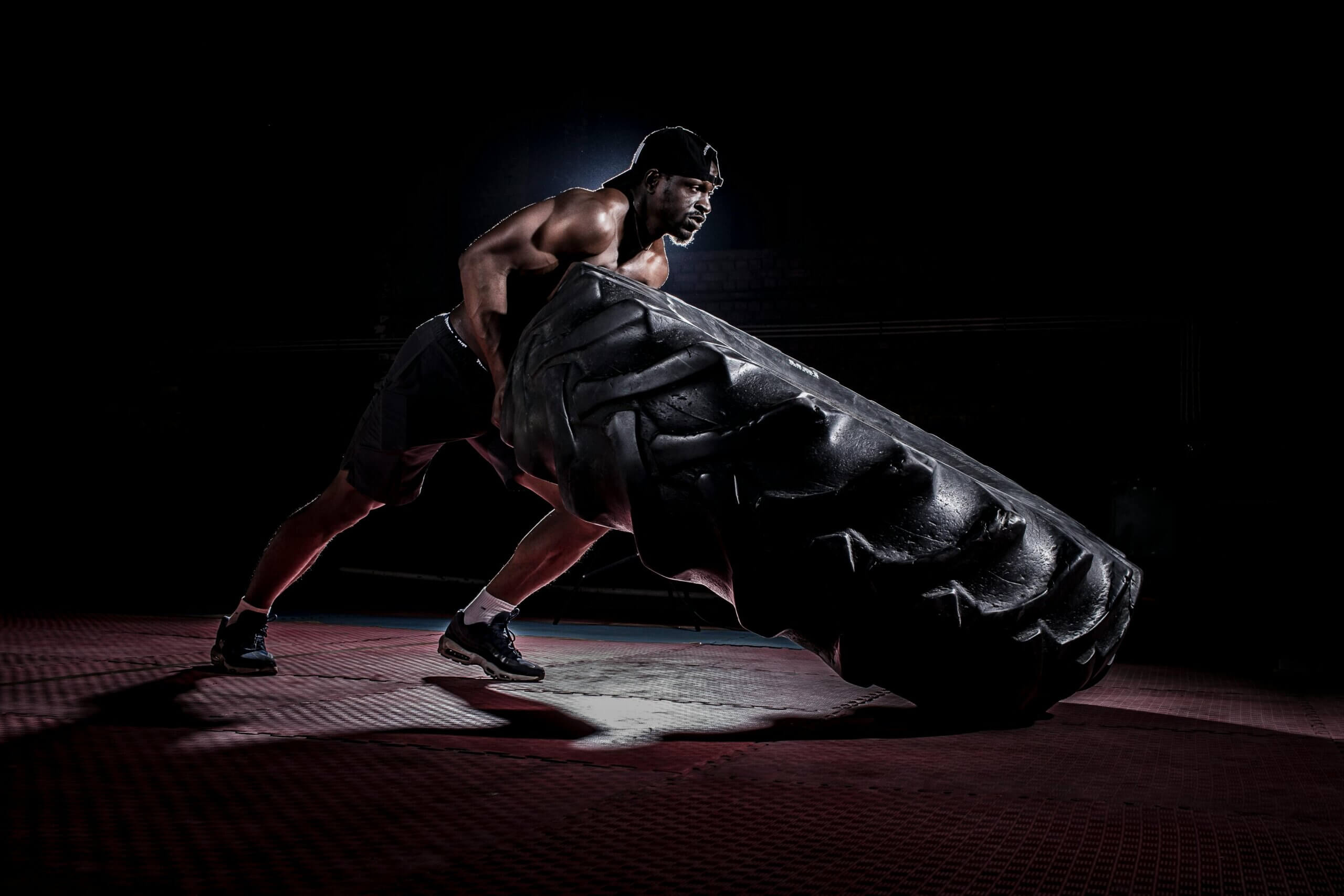 Muscular man wearing black shorts and cap flipping a large tire in a dark gym.