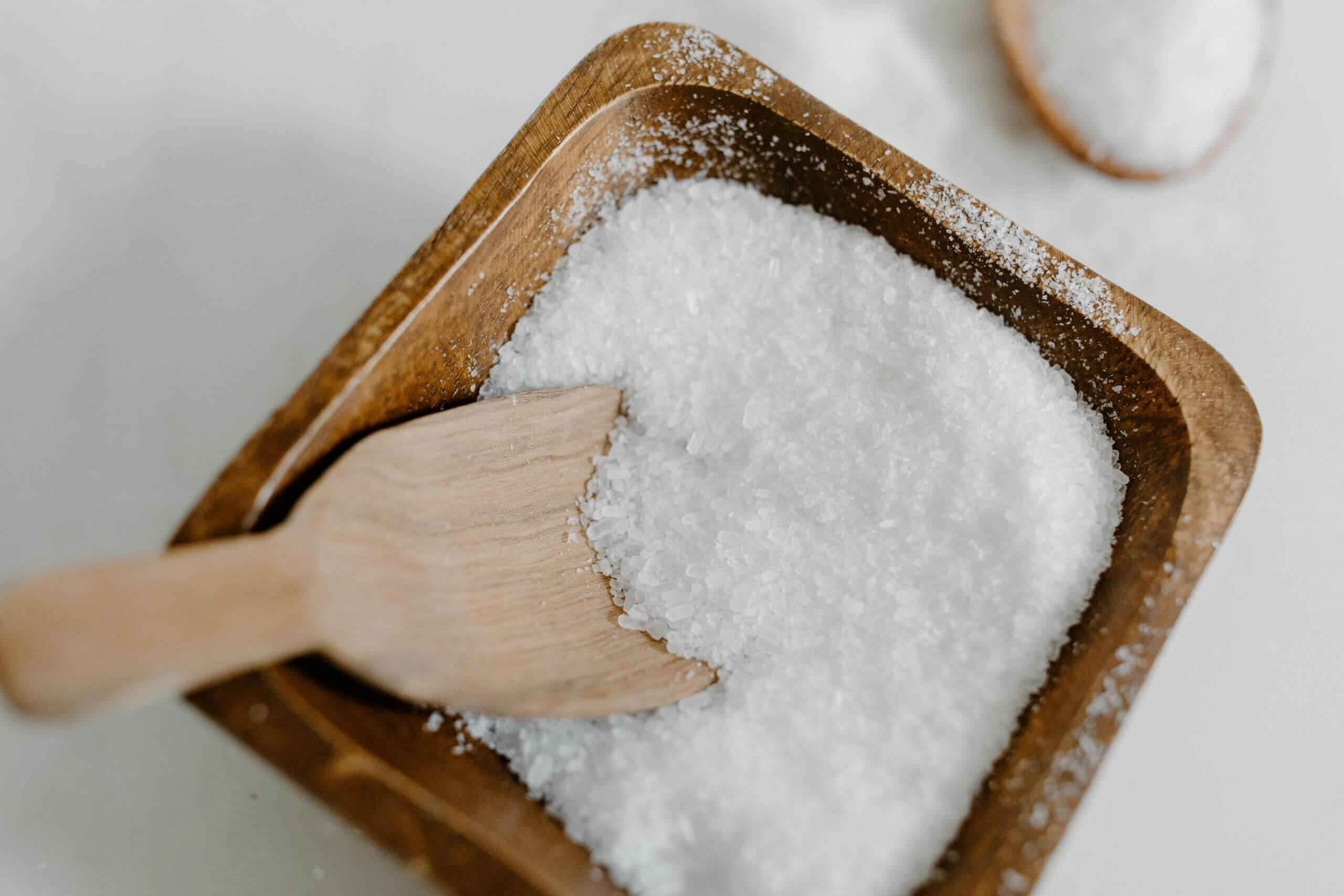 Wooden scoop in a square wooden bowl filled with coarse sea salt on a white surface