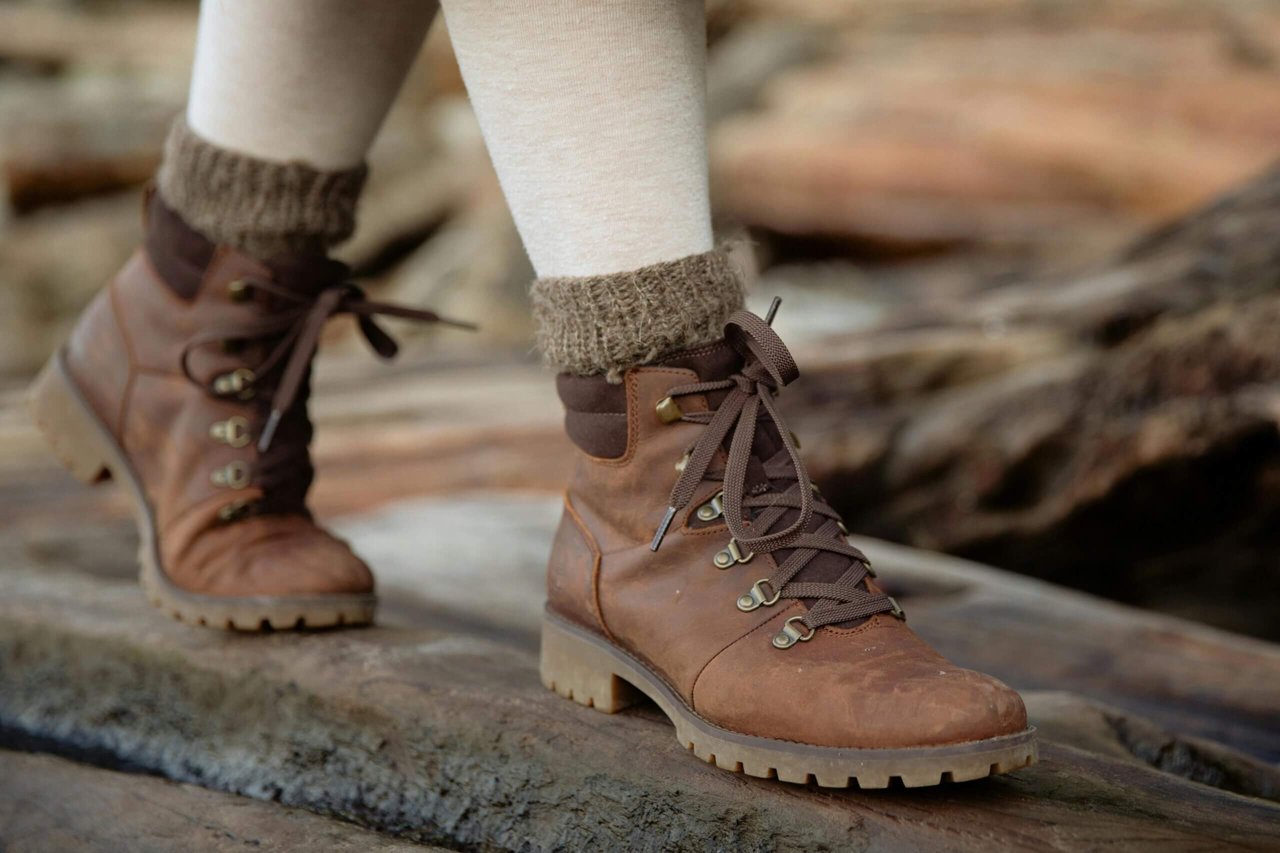Brown leather hiking boots with thick laces worn with beige socks on a wooden surface outdoors.