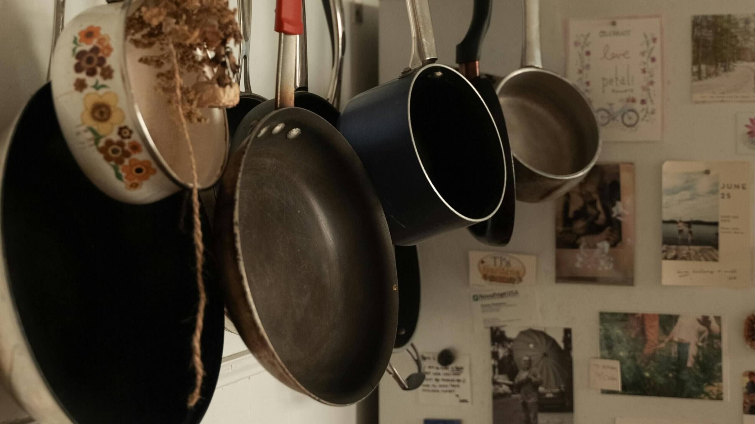 Various frying pans and saucepans hanging on a kitchen wall with photos and notes in the background.