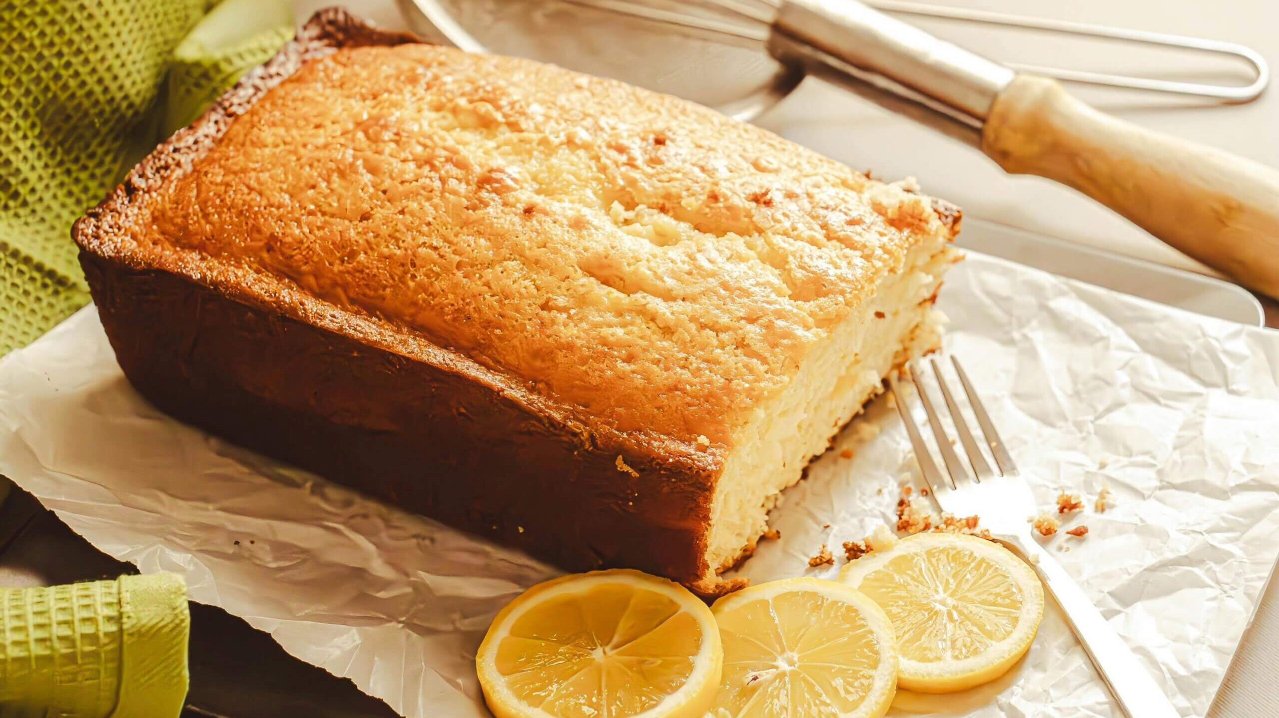 Lemon loaf cake on parchment paper with three lemon slices, a fork, and a whisk in the background