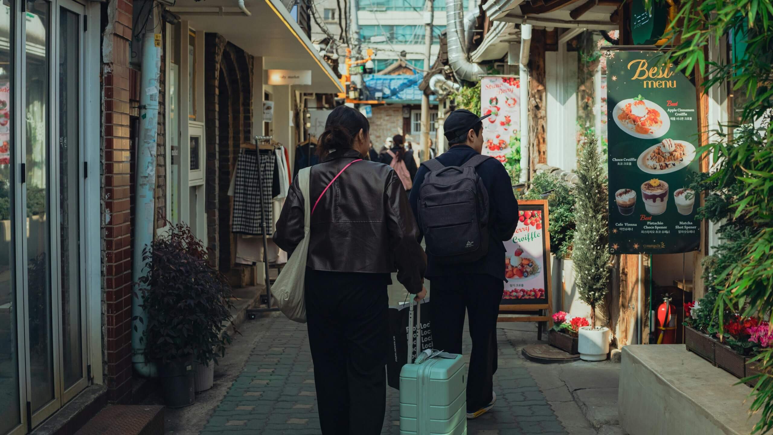 Two people walking down a narrow street with one pulling a mint green suitcase near a cafe menu sign.