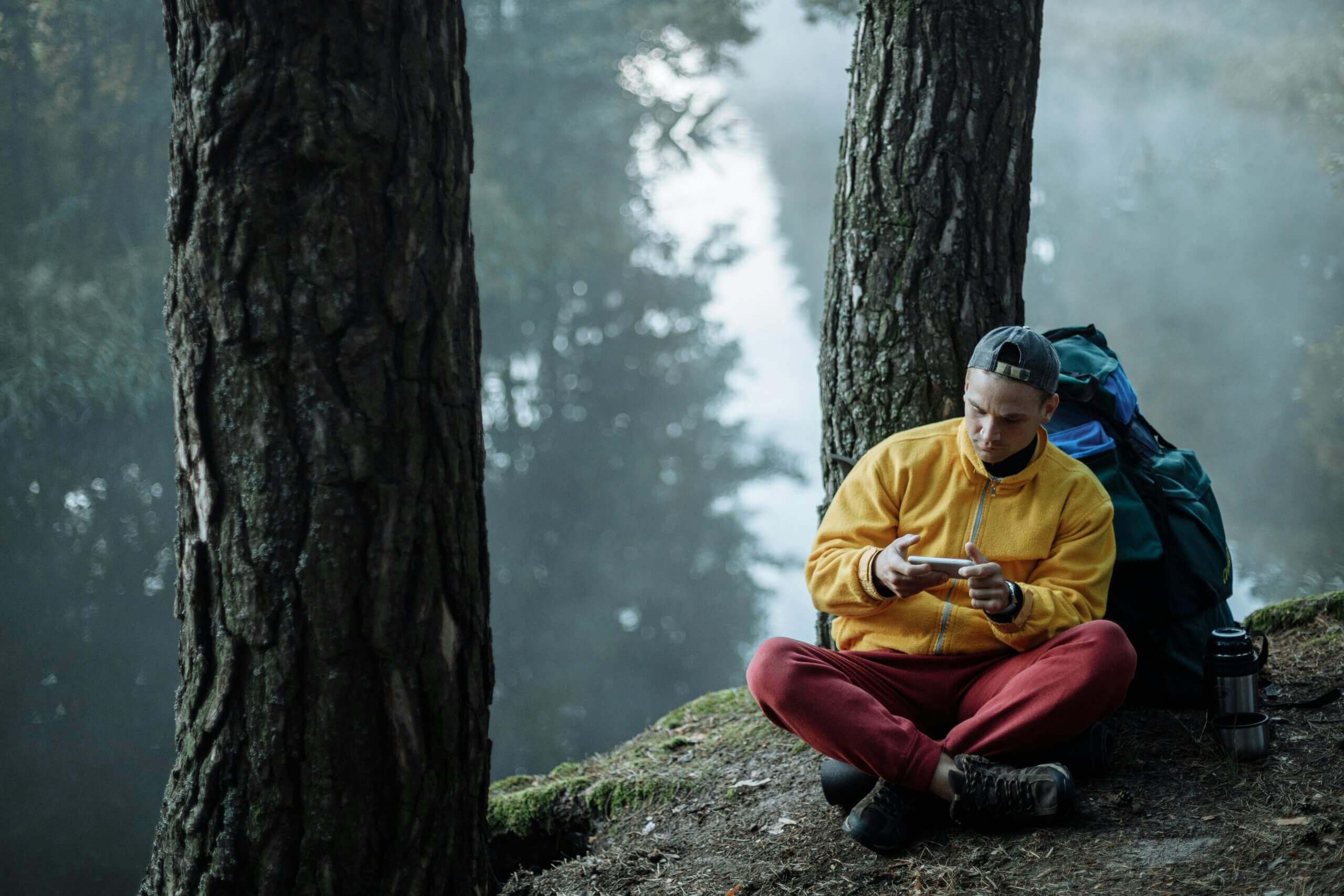 Man in yellow jacket and red pants sitting cross-legged by trees using a smartphone with a backpack and thermos nearby