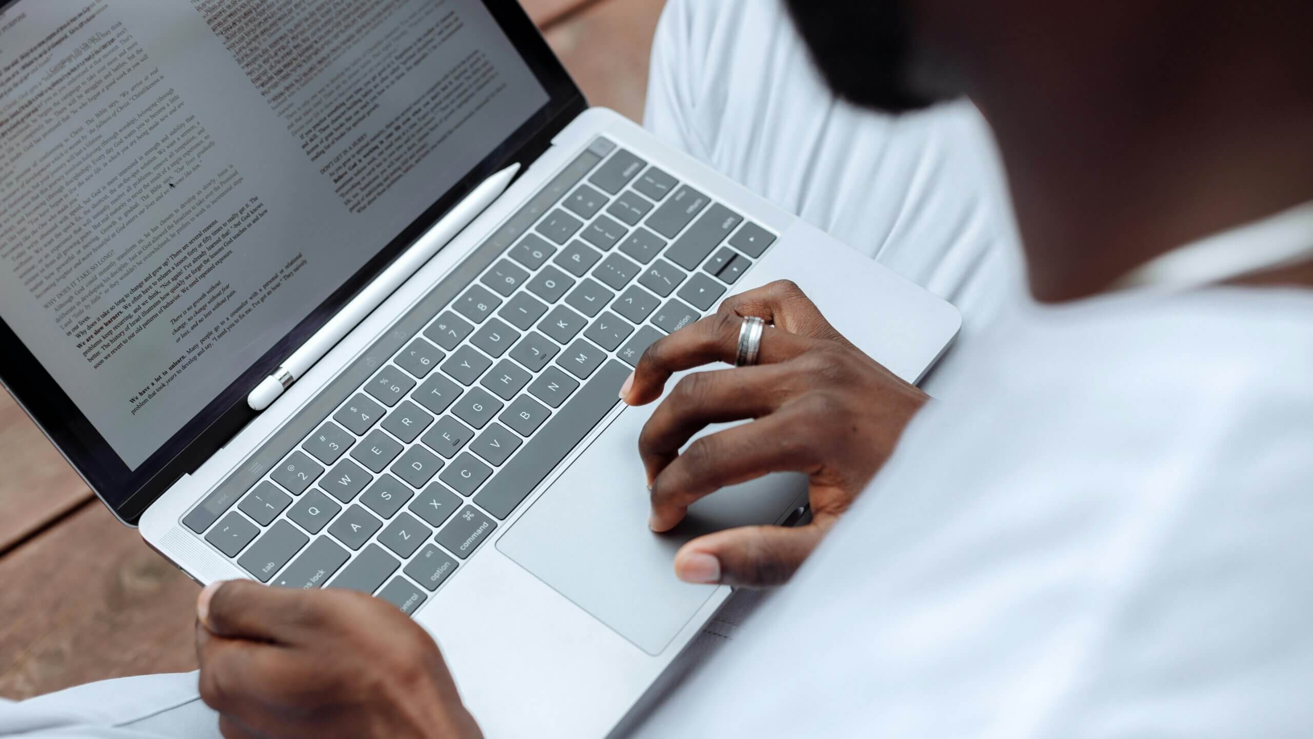 Person using a MacBook laptop with a white stylus resting on the keyboard and reading text on the screen.