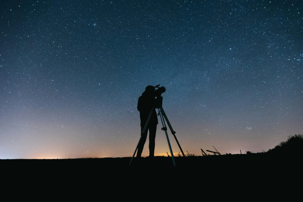 Silhouette of a person using a telescope under a star-filled night sky.