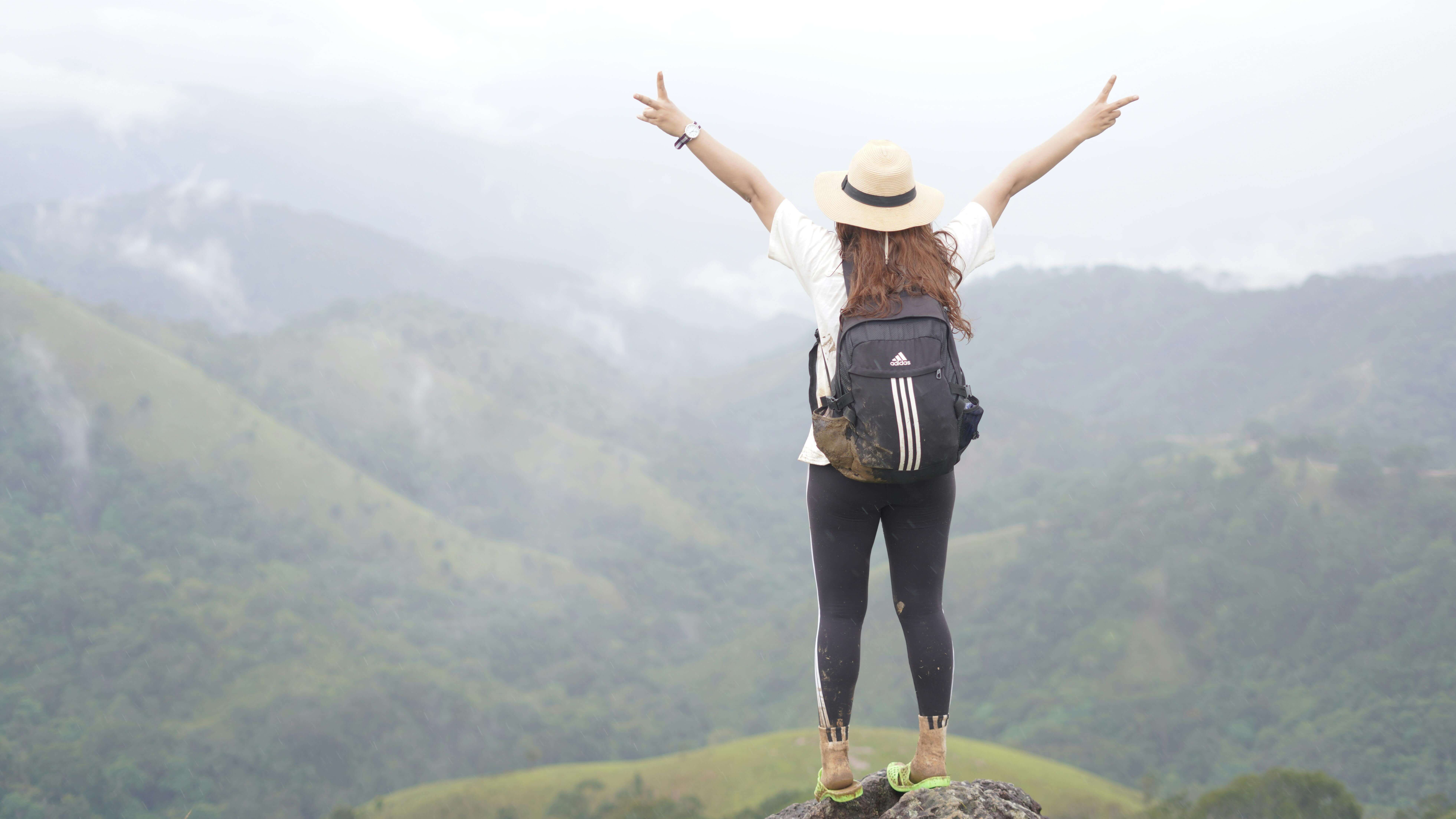 Hiker wearing a straw hat and Adidas backpack standing on a rock with arms raised overlooking misty green hills.