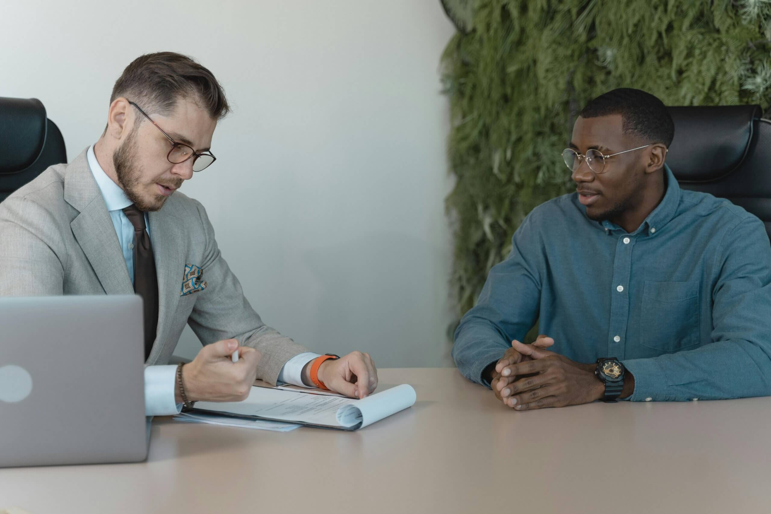 Two men in glasses having a discussion at a desk with documents and a laptop in an office setting.