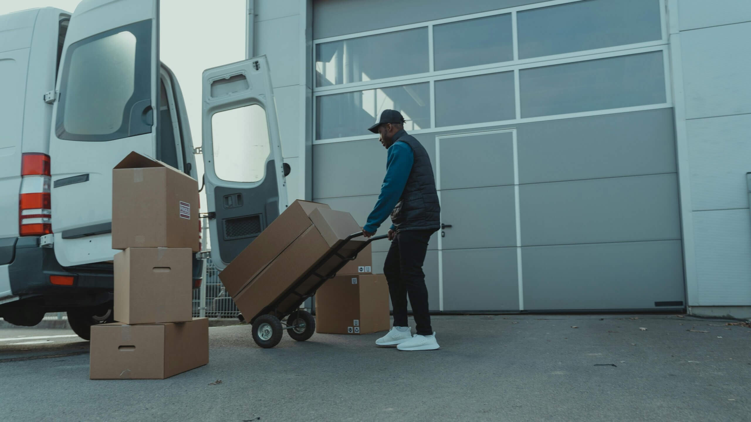 Pria yang mengenakan topi dan rompi membongkar kotak kardus dari van pengiriman berwarna putih dengan truk tangan di luar gudang