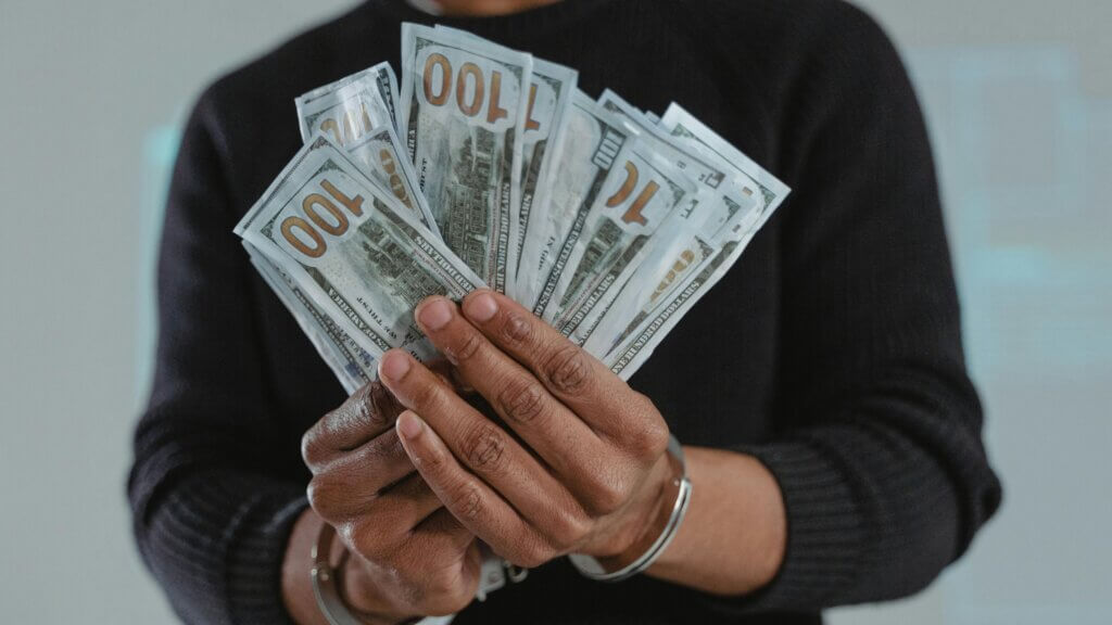 Person holding a fan of US 100 dollar bills wearing a black sweater and silver bracelet