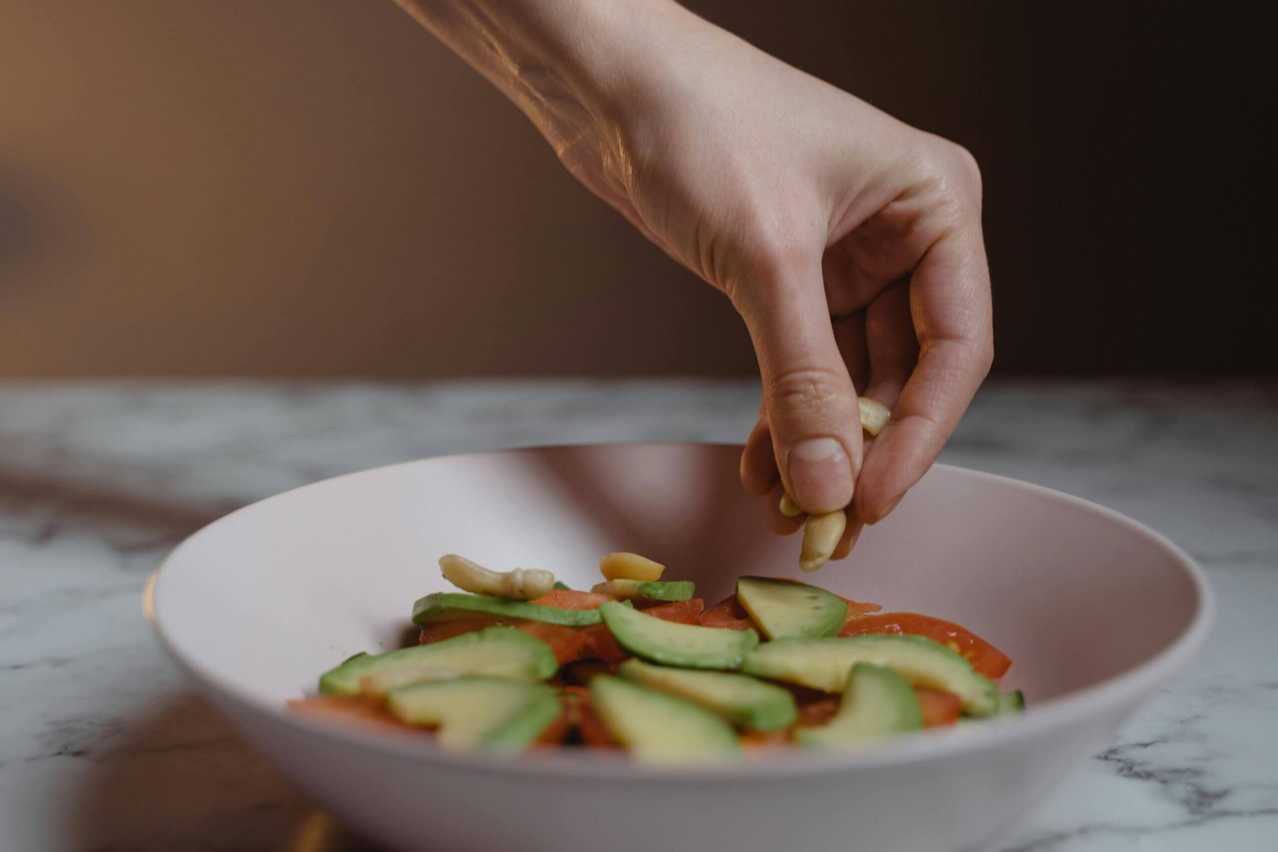 Hand sprinkling cashew nuts onto a salad with avocado and tomato slices in a pink bowl.