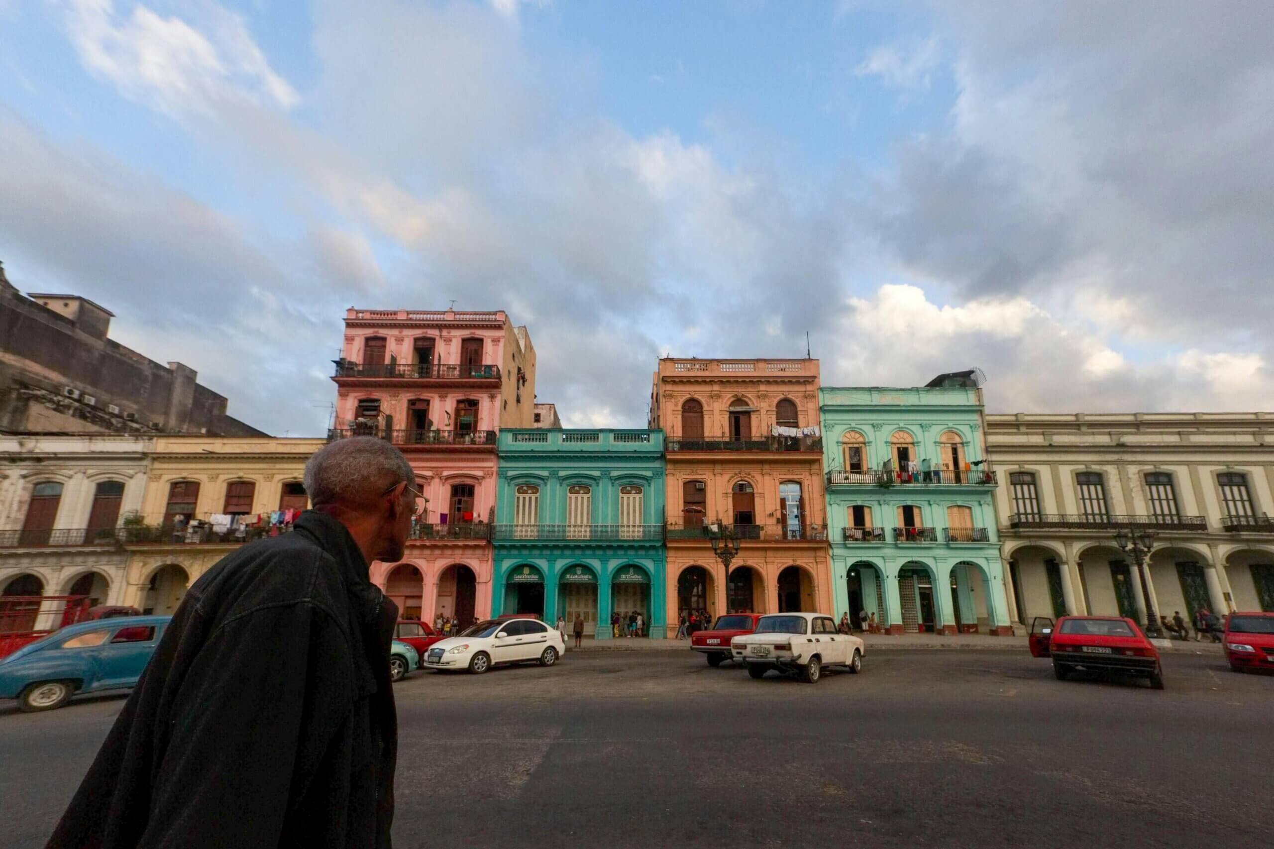 Man in black coat looking at colorful colonial buildings and vintage cars in a city square under a cloudy sky