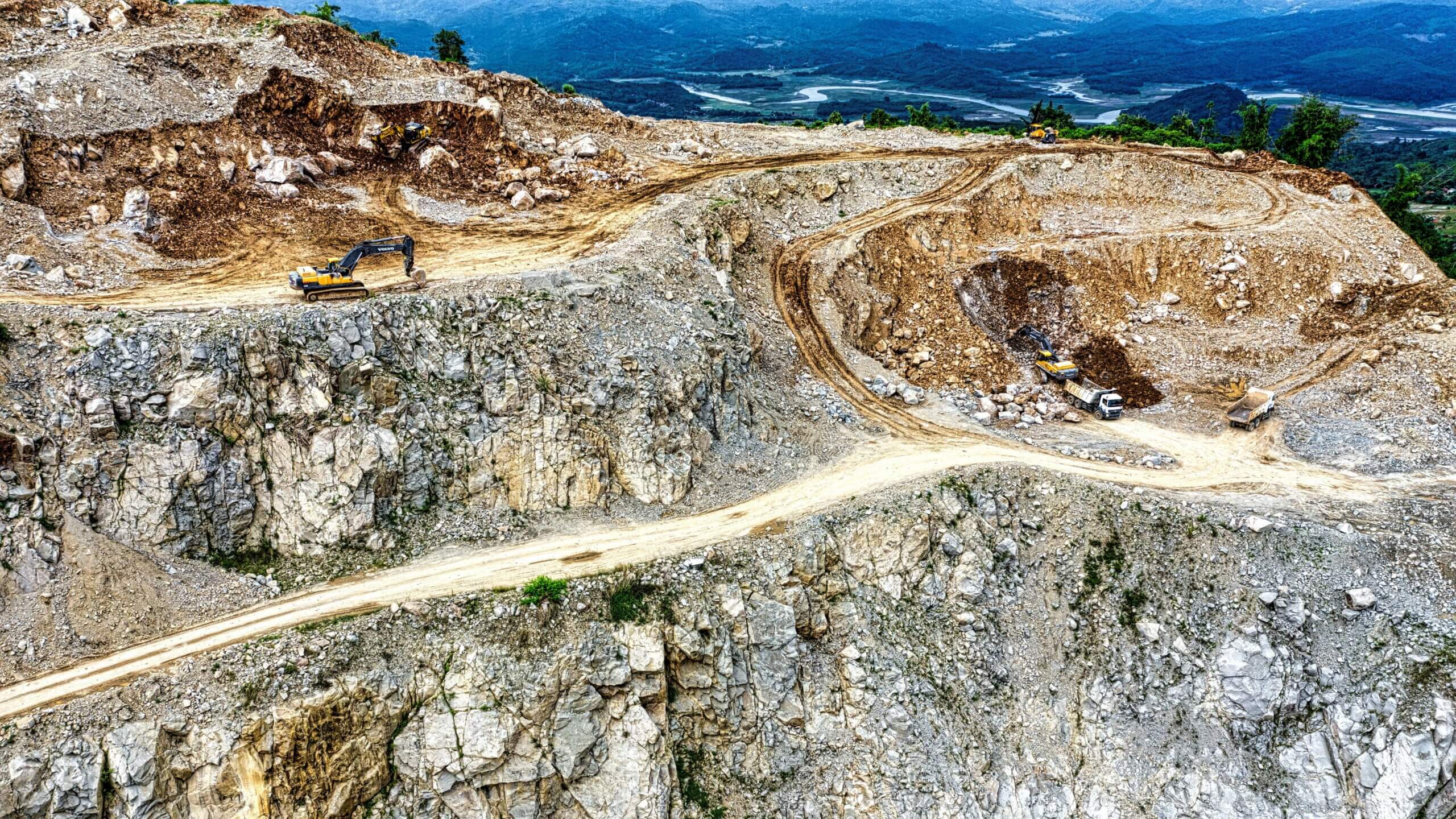Excavators and dump trucks working on a rocky mountain quarry with a river and forest in the background.