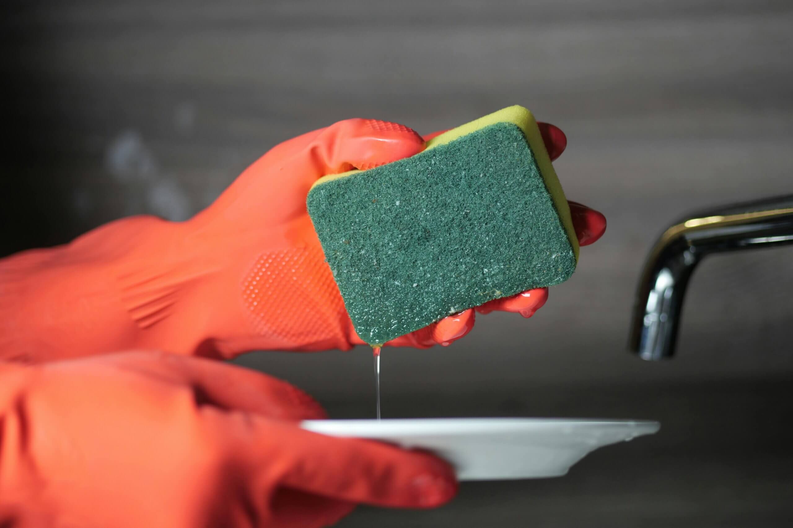 Hand in orange glove holding a wet green and yellow sponge dripping water over a white plate near a faucet