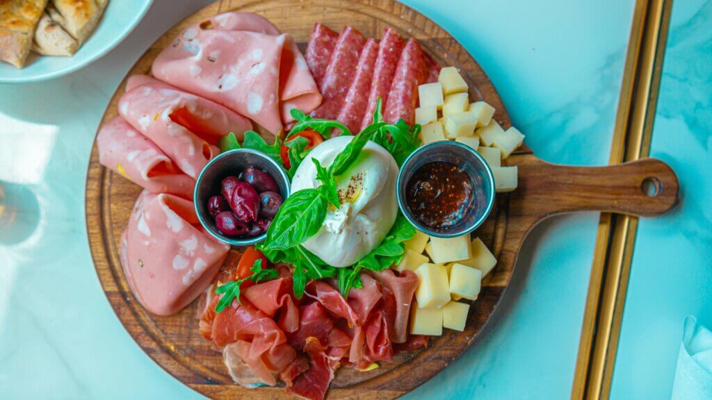 Charcuterie board with sliced meats, cubed cheese, olives, balsamic glaze, and burrata on arugula