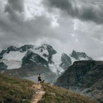 Hiker with a backpack and dog standing on a mountain trail overlooking snow-covered peaks under cloudy sky