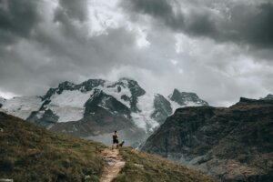 Hiker with a backpack and dog standing on a mountain trail overlooking snow-covered peaks under cloudy sky