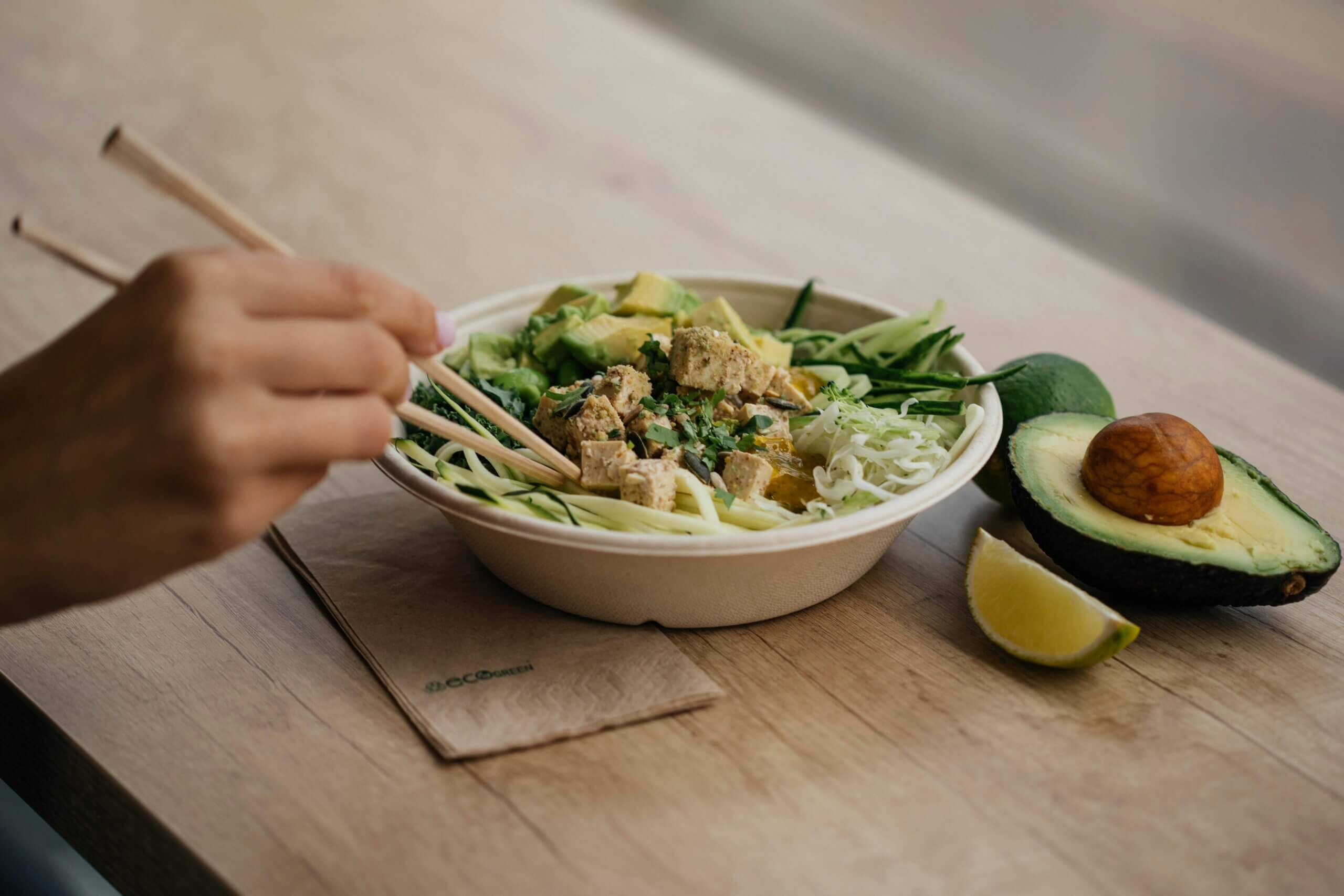 Hand using chopsticks to eat tofu and vegetable salad bowl with avocado and lime wedge on wooden table.