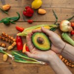 Hands holding half an avocado over a wooden table with assorted fresh vegetables including corn, onions, and peppers.