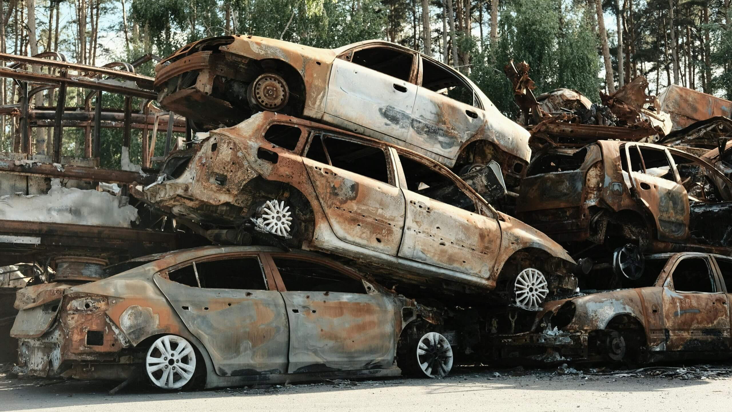 Stack of rusted, burnt-out cars piled in a scrapyard with trees in the background.