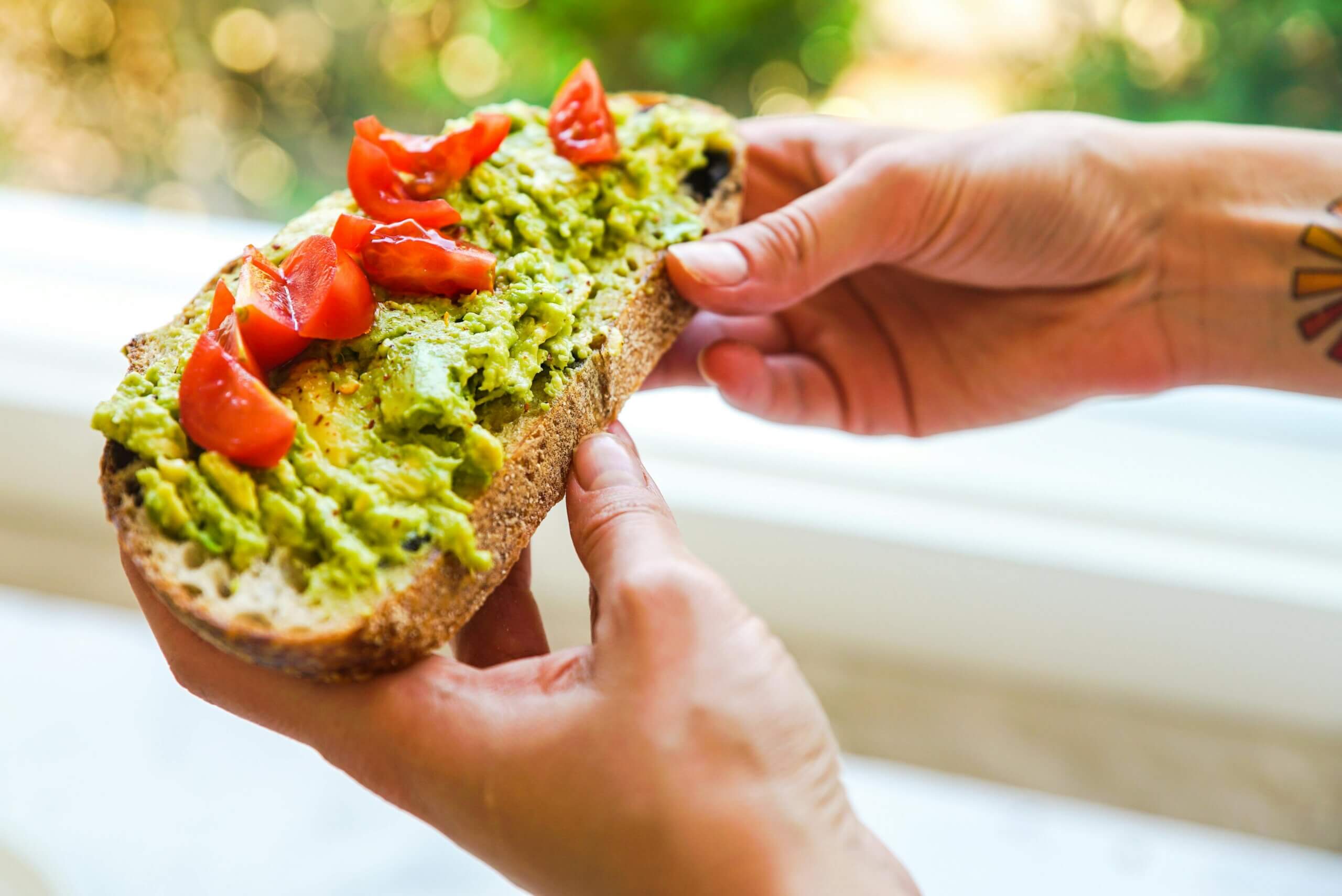 Hands holding a slice of whole grain bread topped with mashed avocado and chopped cherry tomatoes