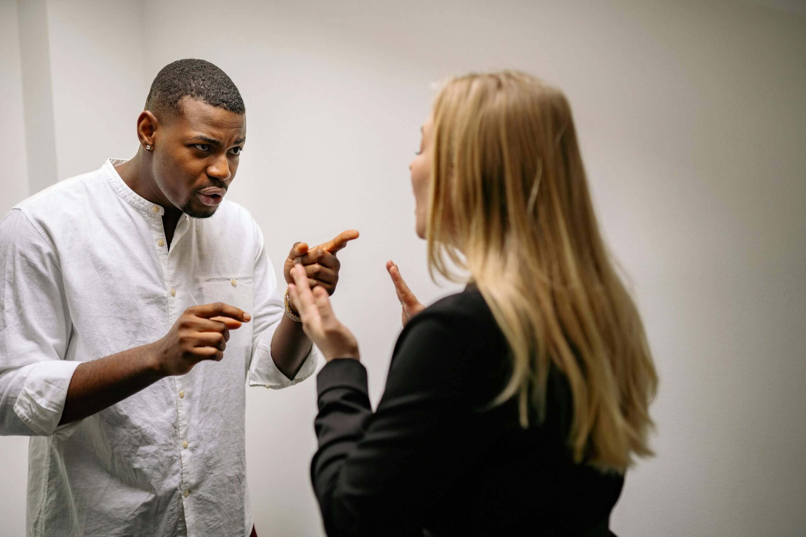 Man in white shirt pointing and arguing with woman in black jacket against plain wall background