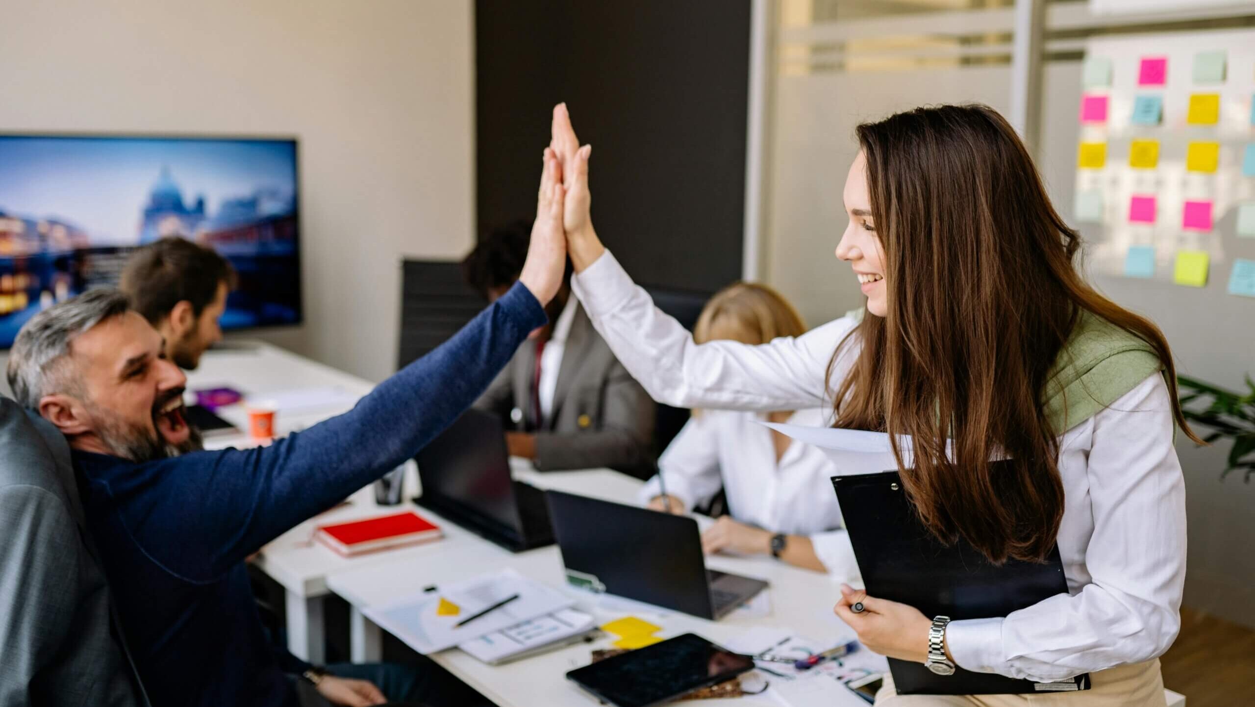 Two coworkers giving a high five in a modern office with laptops and sticky notes on the wall.