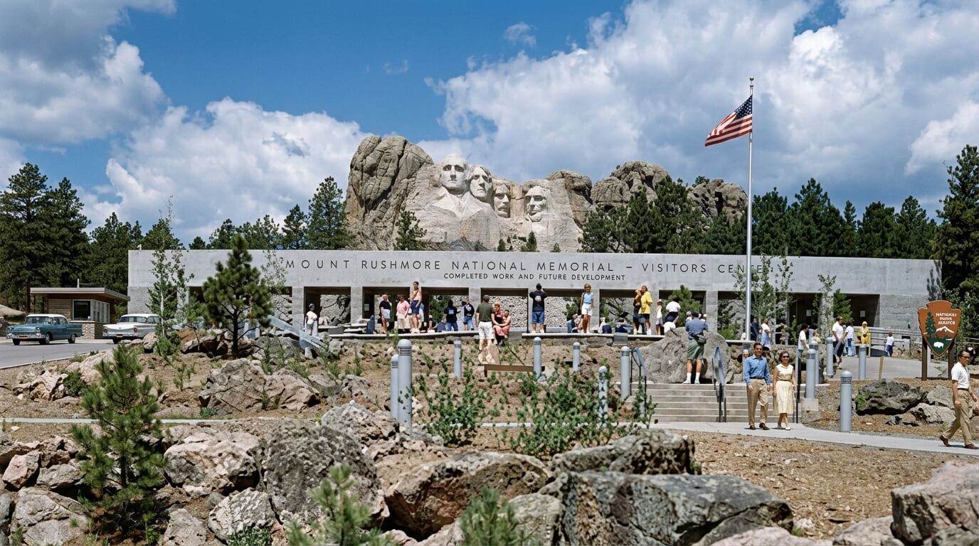 Mount Rushmore National Memorial with visitors and American flag at the entrance under a partly cloudy sky.