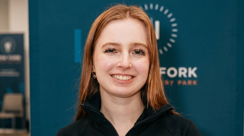 Smiling red-haired woman wearing a black zip-up jacket in front of a blue background with "Fork" text.