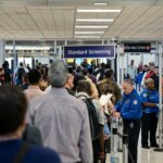 Passengers lined up for TSA standard screening at an airport security checkpoint with officers checking IDs.