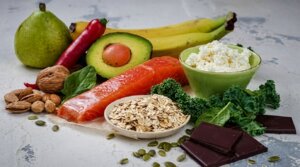 Salmon fillet, avocado, bananas, pear, red chili, nuts, oats, kale, spinach, cottage cheese, and dark chocolate on a white surface.