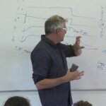 Man in glasses explaining diagrams and notes on a whiteboard during a lecture or presentation