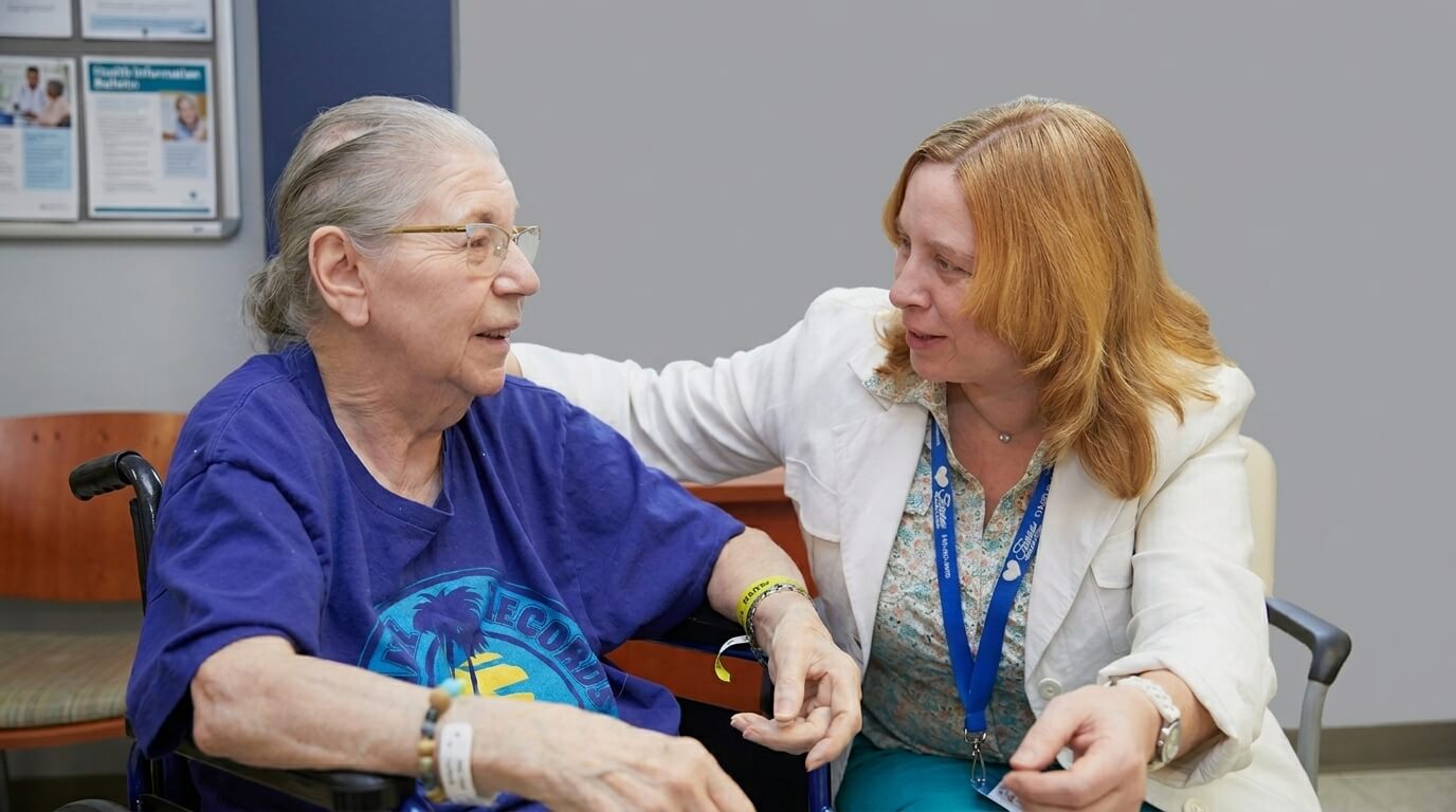 Elderly woman in wheelchair wearing a blue shirt talking with a female healthcare worker in a white coat