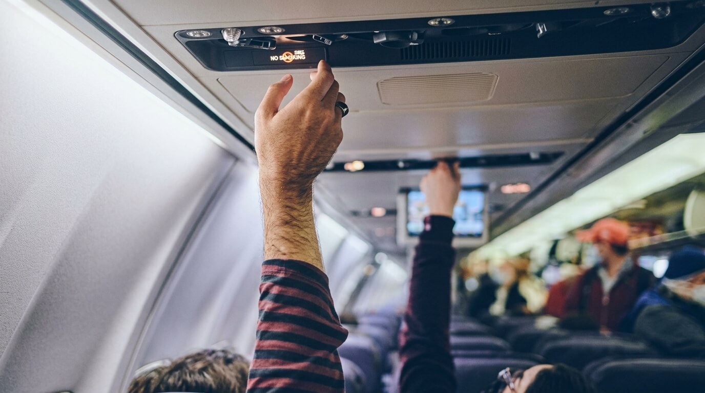 Passenger adjusting overhead air vent and light on an airplane cabin ceiling with other passengers seated.