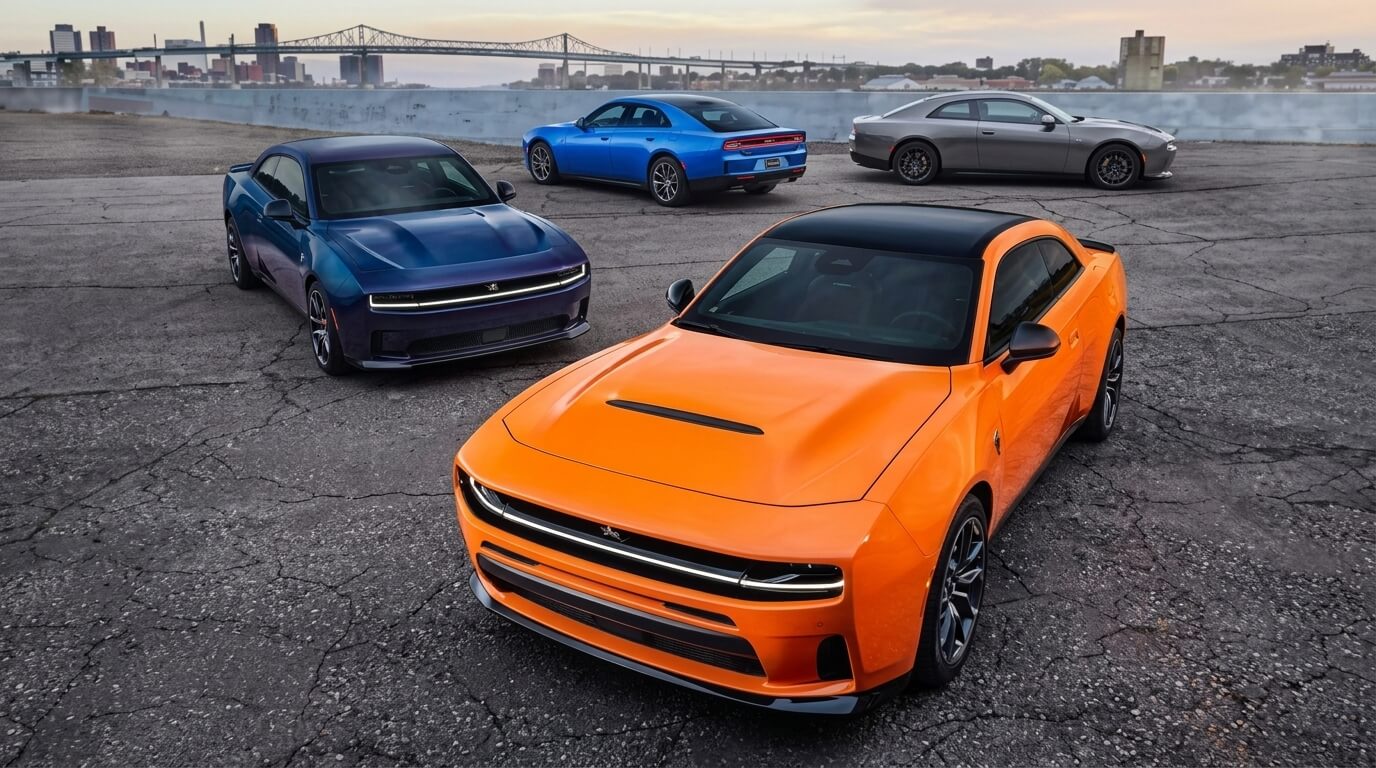 Four Dodge Challenger muscle cars in orange, blue, gray, and dark blue parked on cracked pavement with a city skyline and bridge in the background.