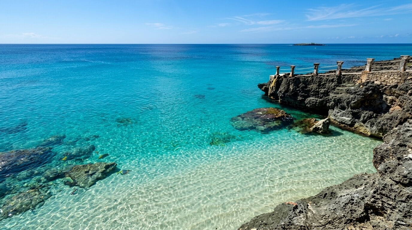 Clear turquoise ocean water with visible rocks and a stone railing on a rocky shoreline under a blue sky